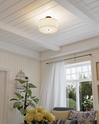 Warm-lit linen drum flush mount installed on a ceiling in a bright room with windows, curtains, and decor