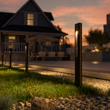 Row of slim black rectangular low-voltage LED path lights light up backyard at sunset with house and patio in the background